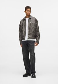 Giubbotto in ecopelle grigia con una leggera lucentezza, chiusura con zip, tasche laterali e dettaglio del logo, abbinato a pantaloni in denim neri e scarpe nere.