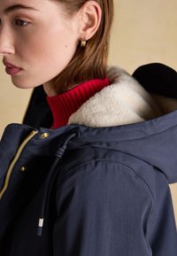 Giovane con orecchini a cerchio dorati, dolcevita rosso a coste e giacca blu navy con cappuccio foderato in pile, rivolto verso sinistra.