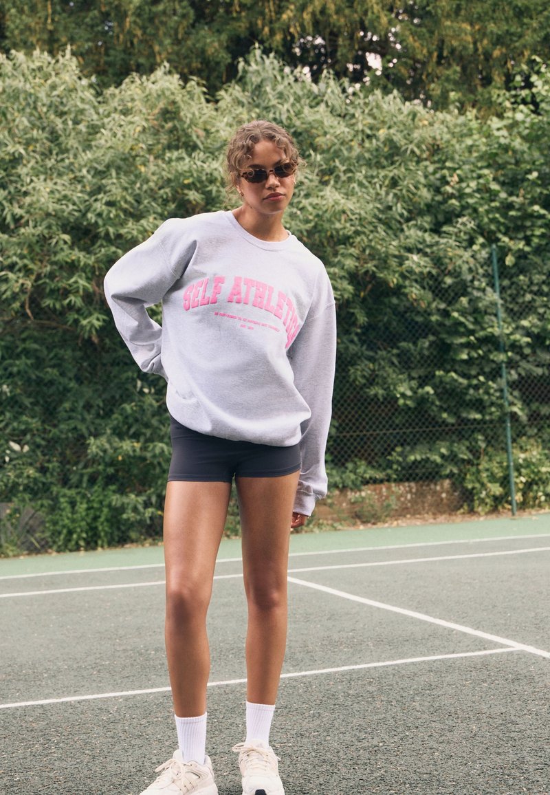 Jong persoon met krullend haar, zonnebril en een oversized grijs sweatshirt, zwarte shorts, witte sokken en sneakers, staand op een tennisbaan.