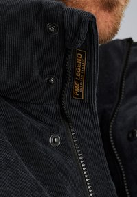 Chaqueta de pana azul marino con cuello alto, botones de presión negros y una etiqueta amarilla con el texto "PME LEGEND" cerca de la cremallera.