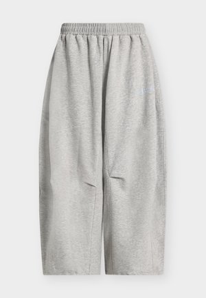 Šviesiai pilkos plačiomis kojomis sportinės kelnės su elastinga juosmens juosta ir subtiliu teksto logotipu viršutinėje šlaunies dalyje.