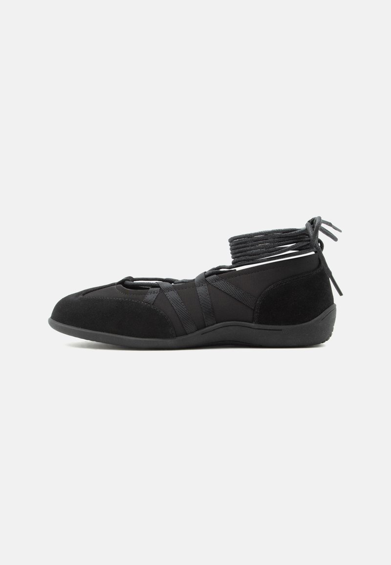 Chaussure plate noire avec brides autour de la cheville et détails en daim, conçue pour un ajustement sûr et un style minimaliste.