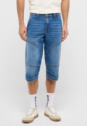 Man wearing knee-length blue denim shorts, white crew socks with text, and beige sneakers, standing against a plain gray background.