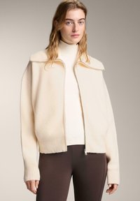 Krém színű polárdzseki zipzáras lezárással, bordázott mandzsettákkal és magas gallérral. Fazonra szabott felső és tompa barna leggings fölött viselve.