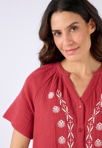 Blusa di cotone rossa con maniche corte, tessuto a trama, scollatura a V e ricami floreali bianchi sulla parte anteriore. Presenta bottoni rosa.