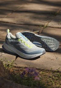 Chaussures de course gris clair et bleu avec des lacets jaune néon reposant sur un chemin de pierre ensoleillé avec de petites plantes à proximité.