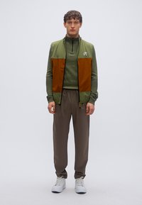 Γιλέκο με μπλοκ χρωμάτων σε πράσινο και καφέ πάνω από πράσινη μπλούζα, συνδυασμένο με φαρδιά καφέ παντελόνια και λευκά αθλητικά παπούτσια. Ομαλή υφή, καθημερινός σχεδιασμός.