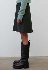 Botas negras hasta la rodilla con suelas gruesas, combinadas con una falda verde oscura hasta la rodilla y un suéter azul claro con mangas de punto texturizado.