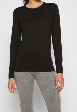 Frau trägt ein enganliegendes schwarzes Langarmshirt und hellgraue Leggings, steht vor einem einfarbigen hellen Hintergrund.