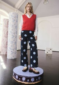 Piros kötött mellény egy fehér ing felett, széles kék farmernadrággal, nagy világoskék pöttyökkel. Barna cipők krémszínű részletekkel.