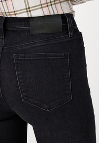Cilvēks valkā tumšus džinsu bikses ar aizmugurējo kabatu un melnu Calvin Klein džinsu etiķeti, savienojumā ar raibajām krekla.