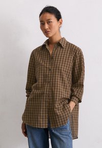 Chemise à carreaux marron et beige avec un col, des manches longues, un boutonnage à l'avant et un ourlet arrière plus long. Portée avec un jean bleu.