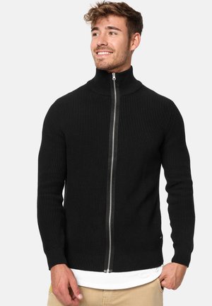 Crna rebrasta zip-up dukserica s visokim ovratnikom, sastojeći se od srebrnog zatvarača i prilagođenih manžeta. Nosena preko bijele košulje.
