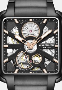 Quadratischer Automatikchronograph mit schwarzem strukturiertem Zifferblatt, silbernen Elementen und goldenen Akzenten. Verfügt über sichtbare Zahnräder und Markierungen an einem metallischen Armband.