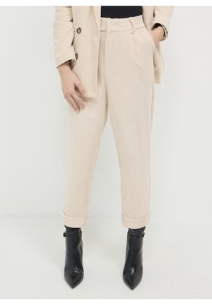 Person trägt beige Cordhosen mit hochgekrempelten Säumen, einen passenden Blazer und schwarze spitze Stiefeletten auf weißem Hintergrund.