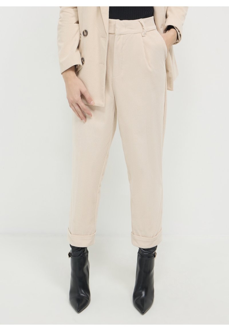 Personne portant un pantalon beige en velours côtelé avec des revers, un blazer assorti et des bottines noires à bout pointu sur fond blanc.