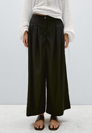 Kvinde iført højtaljede, vidde sorte bukser, hvid tekstureret langærmet sweater og sorte åbne sandaler med tå-ringe, stående på lyst gulv.