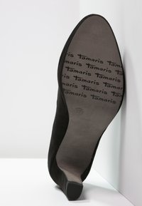 Svart mocka högklackad sko med spetsig tå och avsmalnande klack. Innersulan märkt med upprepande "Tamaris" och storlek "37". Slät textur.