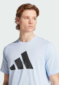 Svetlomodré tričko s krátkym rukávom z mäkkej tkaniny s veľkým čiernym geometrickým logom na prednej strane. Štýl s okrúhlym výstrihom a voľným strihom.