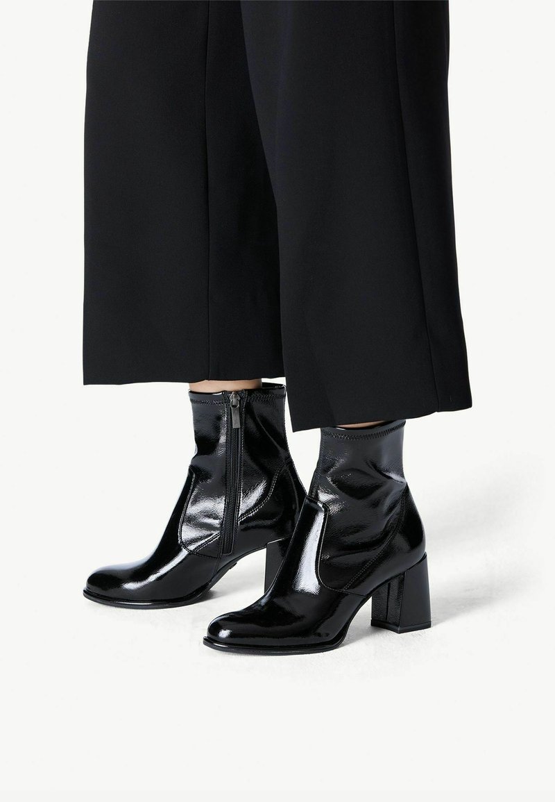 Bottes noires brillantes à chevilles avec talons carrés et fermetures éclair latérales, portées avec un pantalon large noir coupé au-dessus des chevilles.
