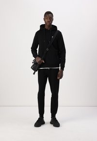 Zwarte hoodie met trekkoord en logo op de borst, gecombineerd met bijpassende afgebogen sweatpants. Draag het met een schoudertas en zwarte sneakers.