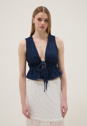 Vrouw met lang blond haar, gekleed in een mouwloos marineblauw topje met een strik aan de voorkant en een witte kanten rok, staand voor een effen achtergrond.