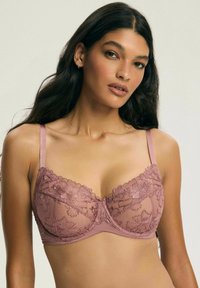 Kobieta z długimi ciemnymi włosami w fioletowym koronkowym biustonoszu z kwiecistym haftem, pozująca na jednolitym jasnym tle.