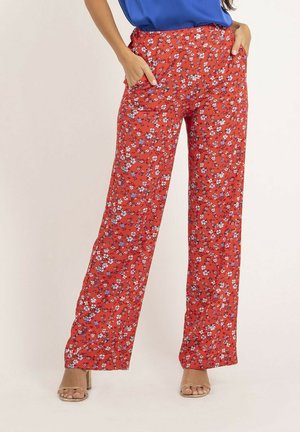 Femme portant un pantalon large fleuri rouge avec poches, associé à un haut bleu et des talons ouverts beige, debout devant un fond uni.