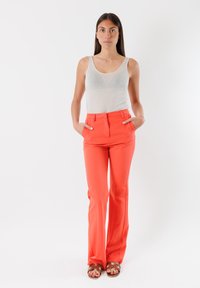 Pantaloni corallo a gamba svasata con una texture liscia e due tasche laterali. Indossati con una canotta grigia chiara e sandali marroni.