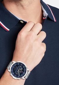 Montre argentée avec un cadran bleu marine, trois sous-cadrans et un bracelet en acier inoxydable ; portée sur une main contre une chemise polo bleu marine avec des accents rouges.
