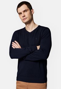 Ciemnoniebieski sweter w serek z miękkiej dzianiny, z długimi rękawami i ściągaczami na mankietach. Dyskretne logo na piersi, w połączeniu z beżowymi spodniami.