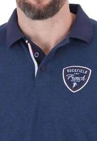 Polo bleu marine avec une finition texturée, présentant un col, une fermeture à boutons et un patch logo sur la poitrine. Comprend des détails d'accent rouge et blanc.