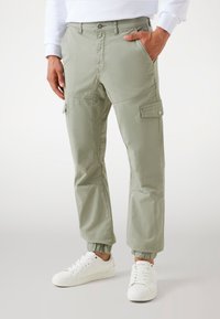 Helle grüne Cargo-Hosen mit elastischen Bündchen, seitlichen Taschen und einer vorderen Knopfverschluss, kombiniert mit weißen Turnschuhen. Glatter, strapazierfähiger Stoff.