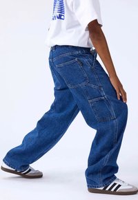 Pantalones cargo de mezclilla azul con piernas anchas, que cuentan con múltiples bolsillos y un ajuste relajado. Combinados con una camiseta blanca y zapatillas gris.