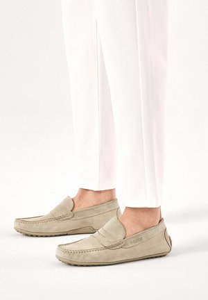Svetlo béžové semišové mokasíny nosené s bielymi nohavicami, zobrazujúce bočný a predný pohľad na topánky s pletenými detailmi.
