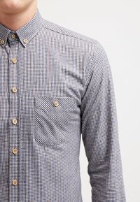 Camicia a maniche lunghe a quadratini con colletto button-down, bottoni in legno e taschino sul petto. I colori sono blu e bianco. Tessuto in cotone.