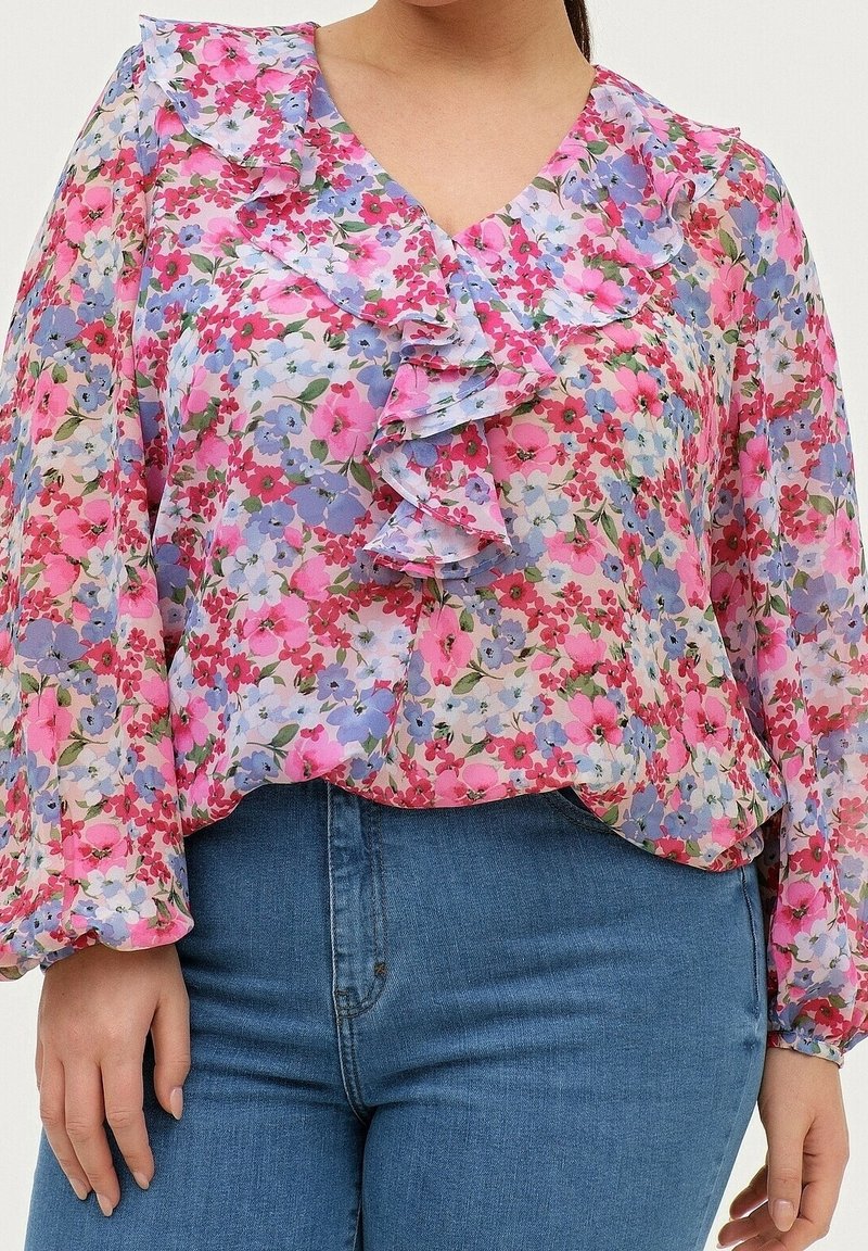 Femme portant un chemisier floral coloré avec un col en V volanté et des manches longues, associé à un jean bleu.