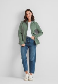 Softshell jacket in olive green, zippered, with a hood and two side pockets. Paired with a white t-shirt and blue straight-leg jeans.