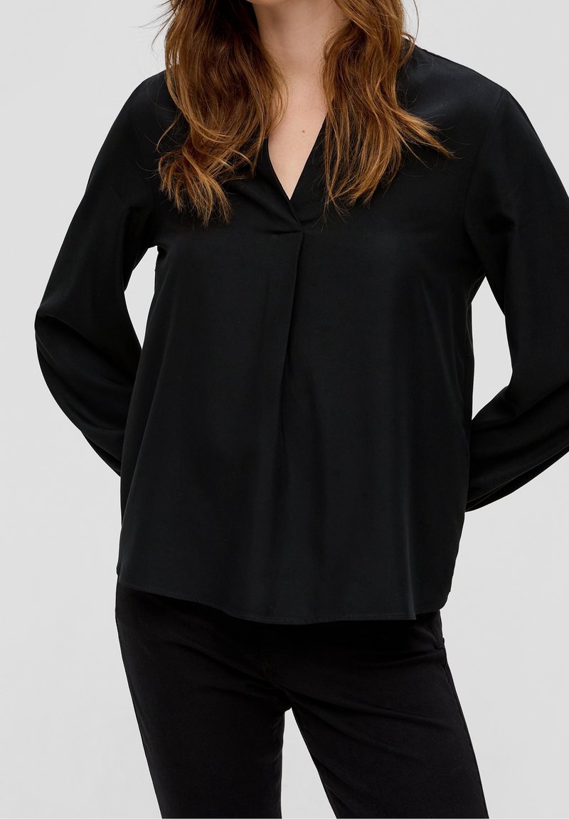 Femme portant un chemisier noir ample à manches longues avec un col en V et un pantalon noir, debout les mains derrière le dos.