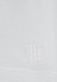 Hvidt stof med subtile broderede initialer "RR" i matchende hvid tråd nær kanten, der viser fin tekstur og syningsdetaljer.