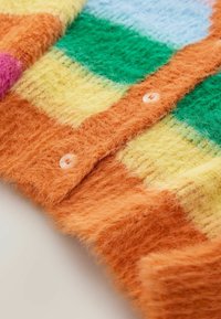 Gros plan sur un cardigan duveteux avec une bande de boutons orange et des blocs multicolores en vert, jaune, orange, bleu et rose.