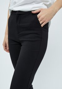 Pantaloni slim-fit neri con una texture liscia, dotati di tasche laterali e vita a media altezza, mostrati con una mano appoggiata sui fianchi.