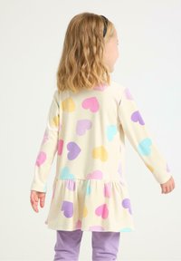 Langærmet kjole i cremefarvet stof med en lagdelt kant, der har flerfarvede hjerte mønstre i pink, lilla, gul og blå.