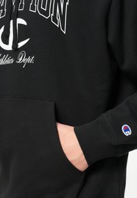 Čierna mikina s kapucňou a predným vreckom. Obsahuje veľký biely grafický text a malé logo Champion na rukáve. Mäkká textúra látky.
