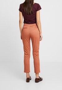 Rust-colored patterned trousers with a fitted design, straight leg, and back pockets, paired with a short-sleeved dark top.