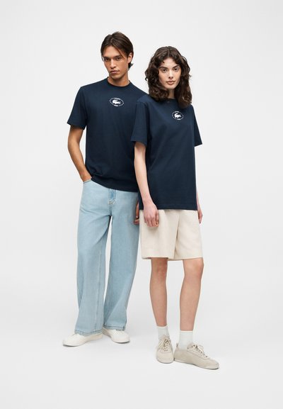 Два модела, носещи тъмносини тениски с логото на Lacoste; единият с светлосини дънки и бели маратонки, а другият с бежови къси панталони и бежови маратонки.