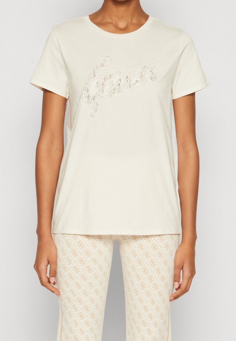 T-shirt crème à manches courtes avec un col rond, orné d'un texte scintillant en script. Tissu doux, coupe décontractée. Porté avec un pantalon à motifs superposé.