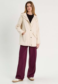 Veste crème clair avec une capuche, dotée de boutons-pression, de poches latérales et d'une coupe décontractée. Associée à un pantalon large violet et des chaussures beiges.