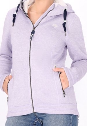 Osoba ubrana w jasnofioletową bluzę z kapturem na zamek z granatowymi sznurkami i rękami w kieszeniach na zamek, w połączeniu z niebieskimi dżinsami.
