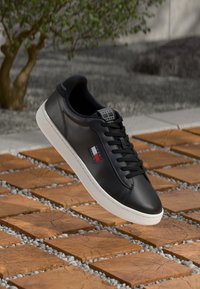 Zwart leren sneaker met witte zool, veters en Tommy Hilfiger-logo zwevend boven houten vierkante tegels met grindgaten buiten.